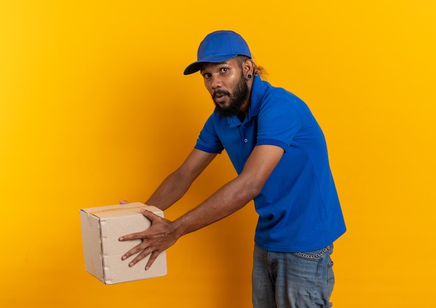 Anxious young afro-american delivery man holding cardboard box isolated on orange background with copy space