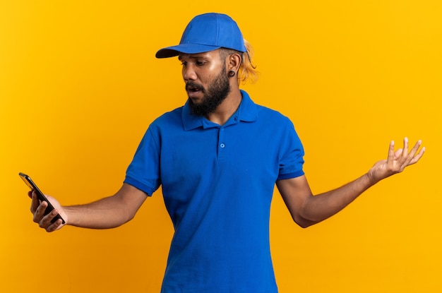 Anxious young afro-american delivery man holding and looking at phone isolated on orange background with copy space