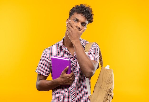 Anxious young afro-american student with backpack holding book and putting hand on his mouth 