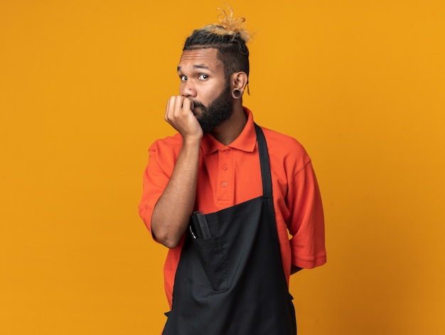 Anxious young barber wearing uniform keeping hand behind back and on lips looking at front isolated on orange wall with copy space