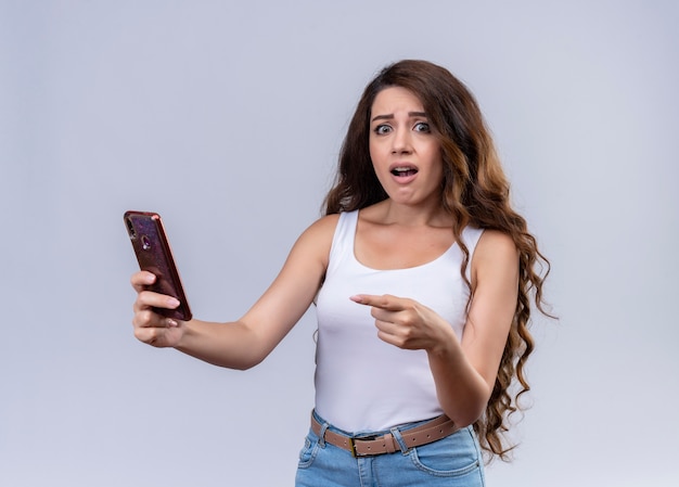 Anxious young beautiful girl holding mobile phone and pointing at it 