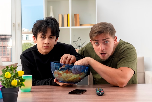 Anxious young blonde and brunette handsome guys sit at table holding and eating bowl of chips inside living room