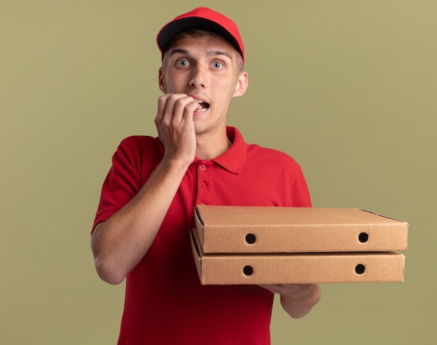 Ansioso giovane biondo consegna ragazzo morde le unghie e tiene scatole per pizza su verde oliva