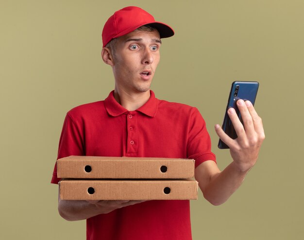 Обеспокоенный молодой блондин курьер держит коробки с пиццей и смотрит на телефон