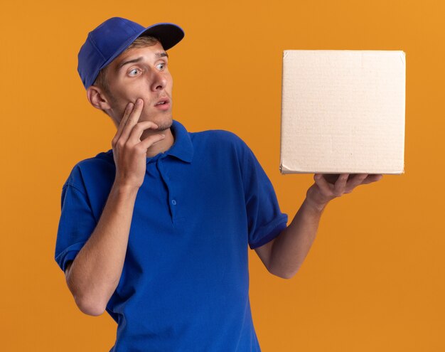 Anxious young blonde delivery boy puts hand on face holding and looking at cardbox isolated on orange wall with copy space