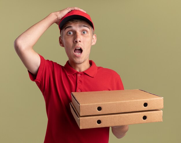 Обеспокоенный молодой блондин-посыльный кладет руку на голову и держит коробки с пиццей на оливково-зеленом