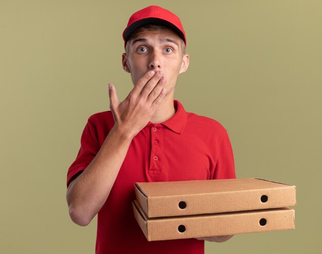 Обеспокоенный молодой блондин-посыльный кладет руку в рот и держит коробки с пиццей на оливково-зеленом