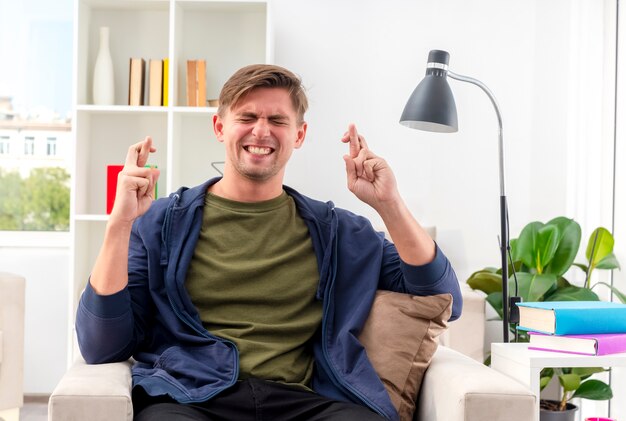 Anxious young blonde handsome man sits on armchair crossing fingers with closed eyes