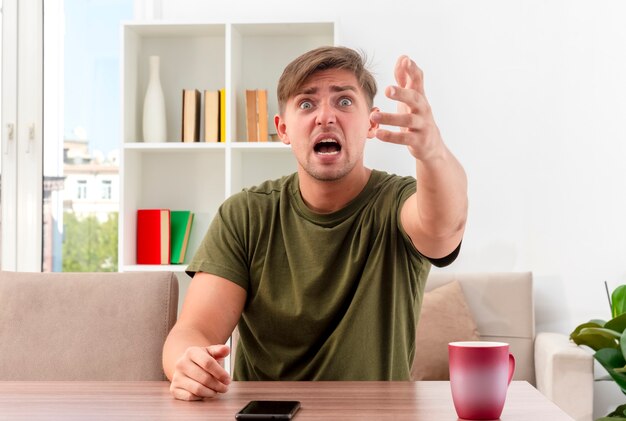 Anxious young blonde handsome man sits at table with cup and phone looking and pointing at camera with hand inside living room