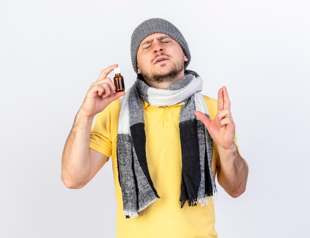 Anxious young blonde ill man wearing winter hat and scarf crosses fingers and holds medicine in glass bottle isolated on white wall
