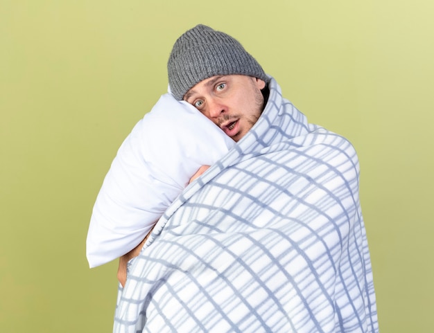 Anxious young blonde ill man wearing winter hat wrapped in plaid hugs pillow isolated on olive green wall