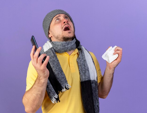 Anxious young blonde ill slavic man wearing winter hat and scarf holds tissue