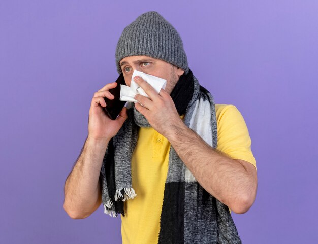 Anxious young blonde ill slavic man wearing winter hat and scarf wipes nose talking on phone