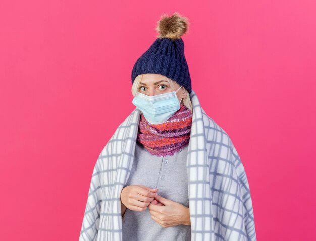 Тревожная молодая блондинка больная славянская женщина в зимней шапке, медицинская маска и шарф, завернутый в плед, держит руки вместе, изолированные на розовой стене с копией пространства