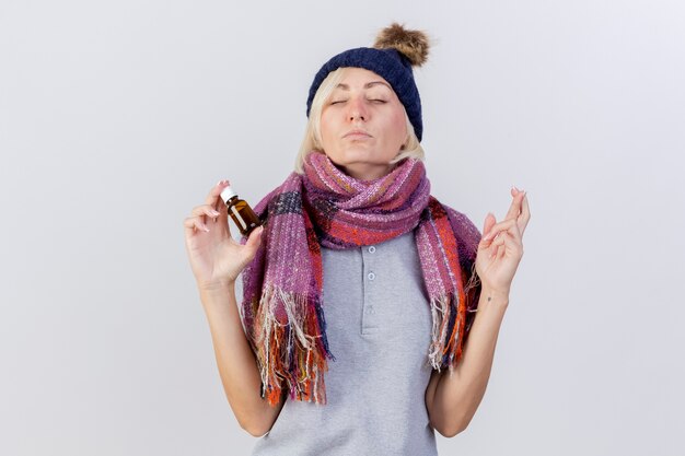 Anxious young blonde ill slavic woman wearing winter hat and scarf crosses fingers and holds medicine in glass bottle isolated on white wall with copy space