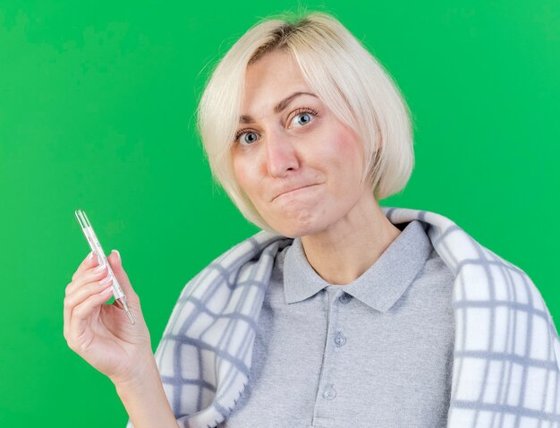 Anxious young blonde ill slavic woman wrapped in plaid holding thermometer isolated on green wall with copy space