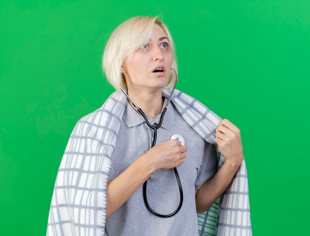 Anxious young blonde ill slavic woman wrapped in plaid holds stethoscope isolated on green wall with copy space