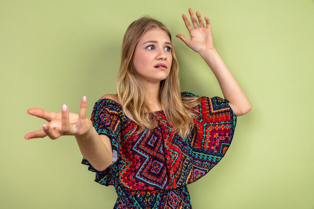 Anxious young blonde slavic girl standing with raised hands and looking at side 