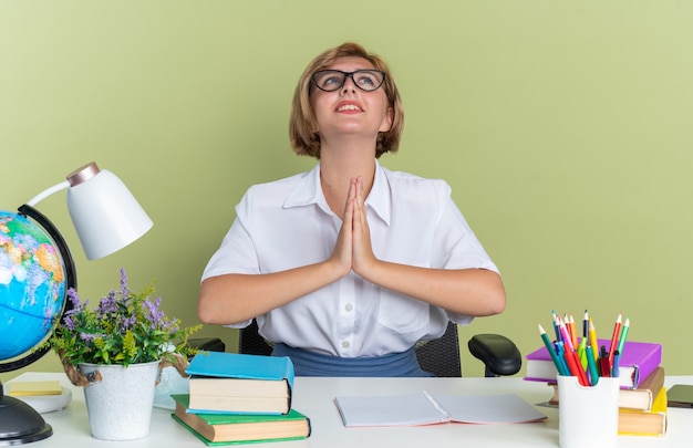 Foto gratuita ansiosa giovane studentessa bionda con gli occhiali seduto alla scrivania con gli strumenti della scuola alzando lo sguardo tenendo le mani insieme pregando isolato sul muro verde oliva