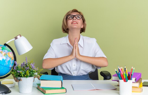 Anxious young blonde student girl wearing glasses sitting at desk with school tools looking up keeping hands together praying isolated on olive green wall