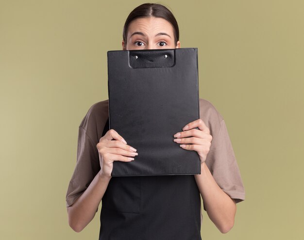 Anxious young brunette barber girl in uniform holds clipboard in front of face on olive green