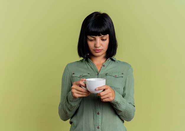 Anxious young brunette caucasian girl holds and looks at cup 