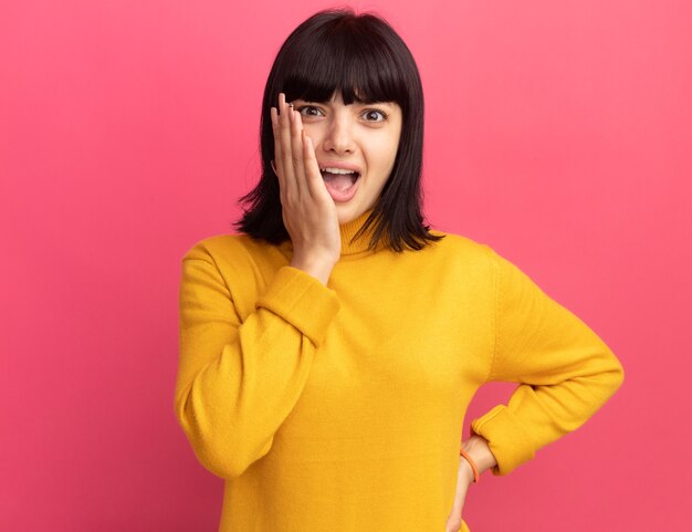 Anxious young brunette caucasian girl puts hand on face and looks at camera on pink