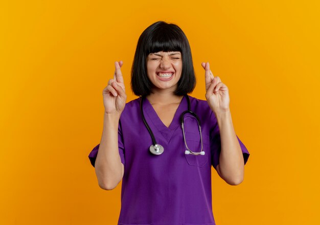 Anxious young brunette female doctor in uniform with stethoscope crossing fingers with closed eyes isolated on orange background with copy space