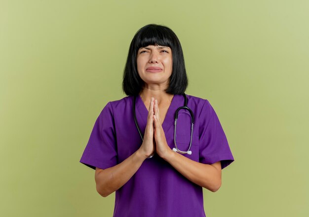 Anxious young brunette female doctor in uniform with stethoscope holds hands together looking up 