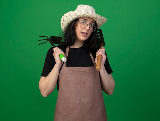 Anxious young brunette female gardener in optical glasses and uniform wearing gardening hat holds rake and hoe rake isolated on green wall