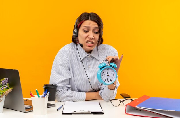 Anxious young call center girl wearing headset sitting at desk holding and looking at alarm clock isolated on orange 