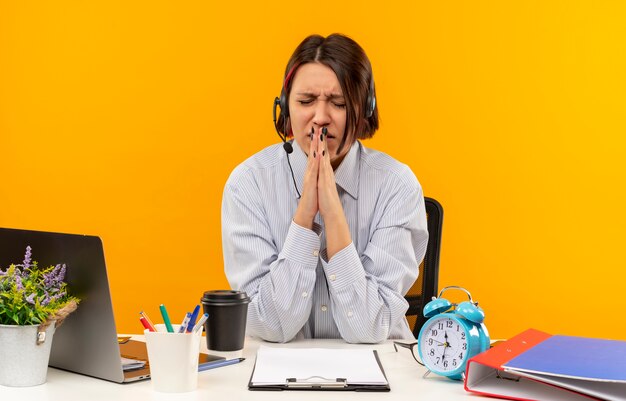 Anxious young call center girl wearing headset sitting at desk putting hands in pray gesture with closed eyes isolated on orange 