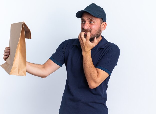 Anxious young caucasian delivery man in blue uniform and cap stretching out paper package looking at it keeping hand in front of chin isolated on white wall
