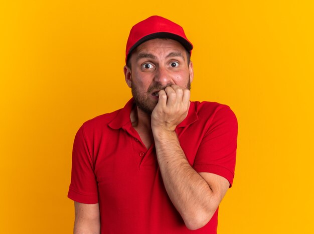 Anxious young caucasian delivery man in red uniform and cap biting fingers looking at camera isolated on orange wall