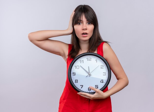 anxious young caucasian girl holding clock and holding head looking at camera on isolated white background