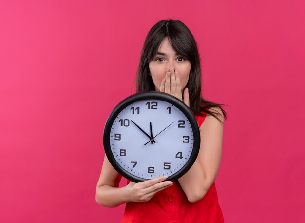 Anxious young caucasian girl holding clock and holding mouth looking at camera on isolated pink background