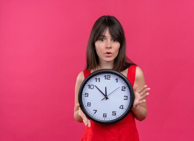 Anxious young caucasian girl holding clock with both hands and looking at camera on isolated pink background with copy space