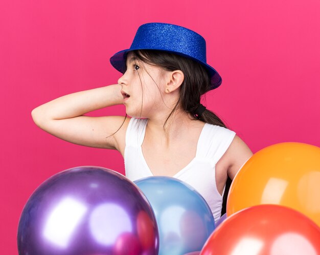 anxious young caucasian girl wearing blue party hat standing with helium balloons putting hand on head and looking at side isolated on pink wall with copy space