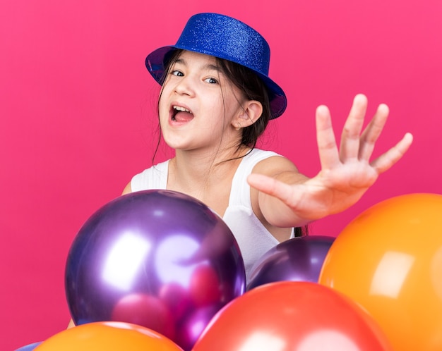 anxious young caucasian girl wearing blue party hat standing with helium balloons and stretching out hand isolated on pink wall with copy space