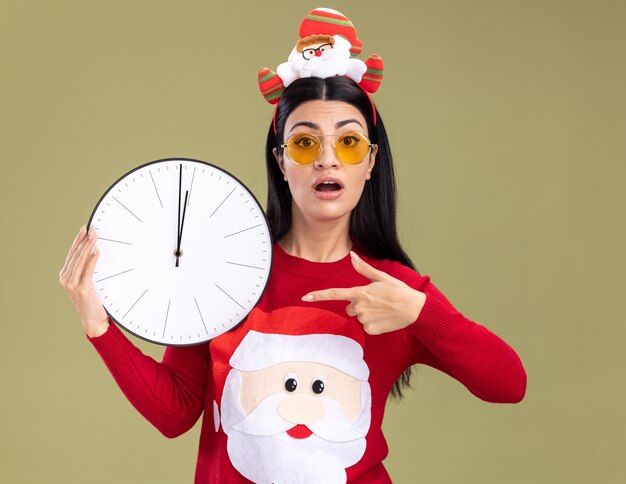 Ansiosa giovane ragazza caucasica che indossa la fascia di babbo natale e maglione con gli occhiali che tiene e che indica l'orologio che guarda l'obbiettivo isolato su priorità bassa verde oliva