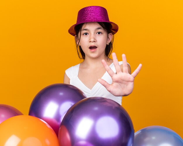 встревоженная молодая кавказская девушка с фиолетовой шляпой жестикулирует знак стоп рука, стоящая с гелиевыми шарами, изолированными на оранжевой стене с копией пространства