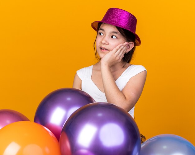 anxious young caucasian girl with purple party hat putting hand on face and looking at side standing with helium balloons isolated on orange wall with copy space