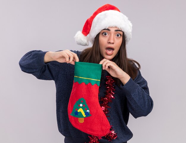 Anxious young caucasian girl with santa hat and garland around neck holds christmas stocking 