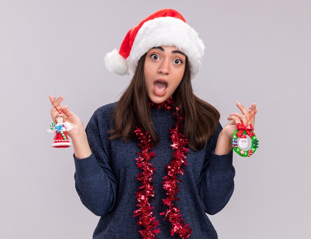 首の周りにサンタの帽子と花輪を持つ気になる若い白人の女の子は、クリスマスツリーのおもちゃを保持します