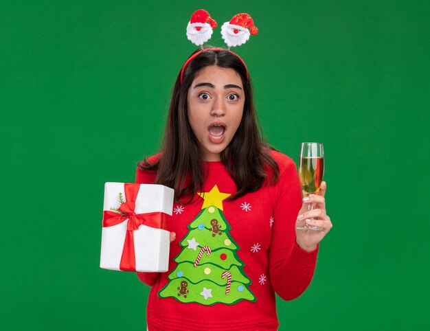 サンタのヘッドバンドを持つ気になる若い白人の女の子は、コピースペースで緑の背景に分離されたシャンパンとクリスマスギフトボックスのガラスを保持します。