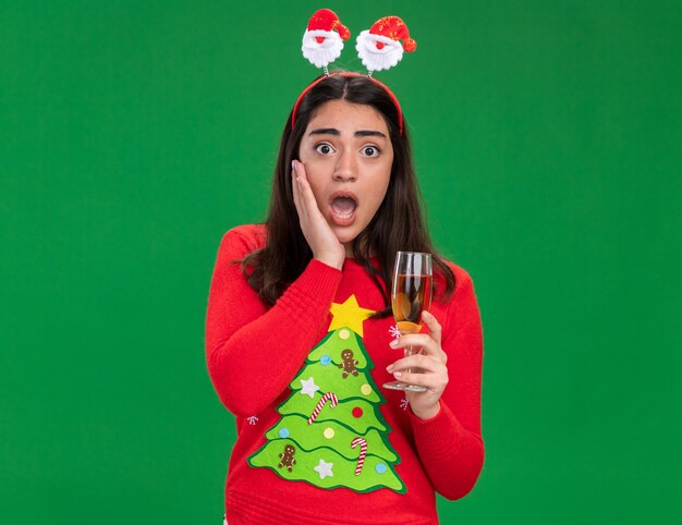 anxious young caucasian girl with santa headband puts hand on face and holds glass of champagne isolated on green background with copy space