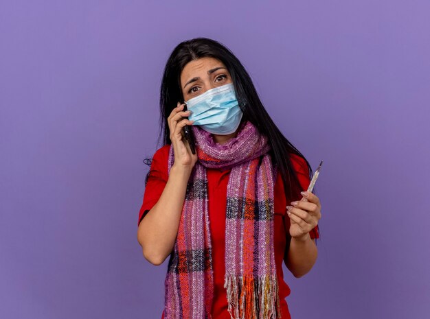 Anxious young caucasian ill girl wearing mask and scarf holding thermometer  isolated on purple wall with copy space