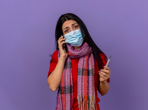 Anxious young caucasian ill girl wearing mask and scarf holding thermometer  isolated on purple wall with copy space