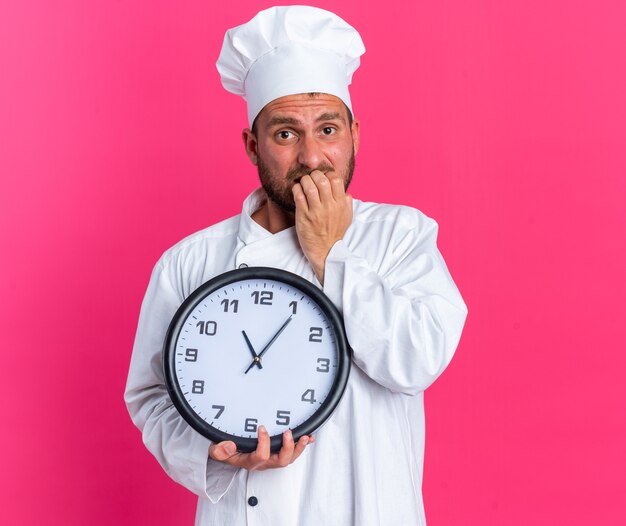 Anxious young caucasian male cook in chef uniform and cap holding clock looking at camera biting fingers isolated on pink wall