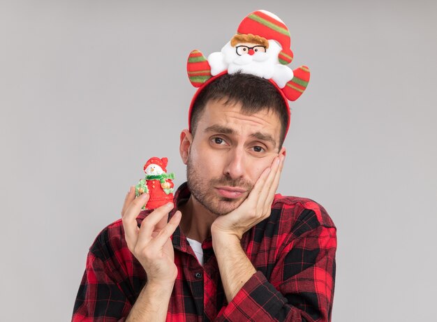 Anxious young caucasian man wearing santa claus headband holding snowman christmas ornament  keeping hand on face isolated on white wall